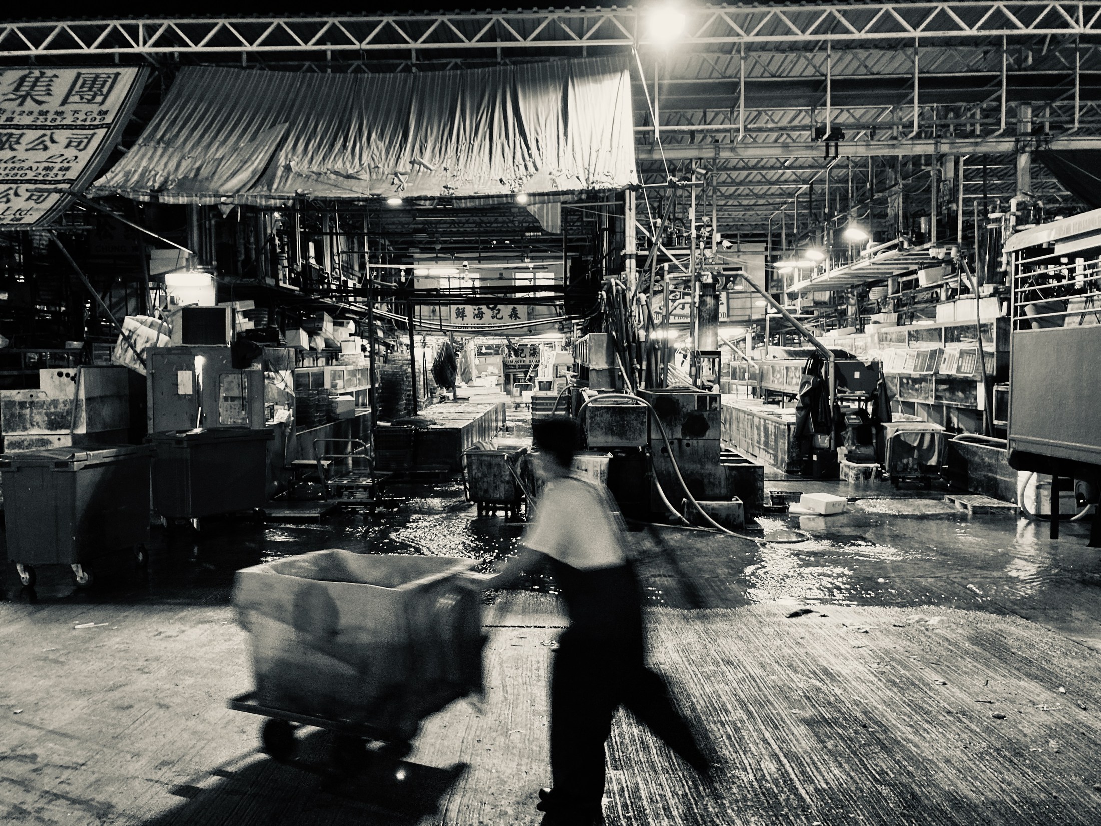 A blurred worker pushes a loaded cart through a wet market hall beneath a dense lattice of lights and pipes.