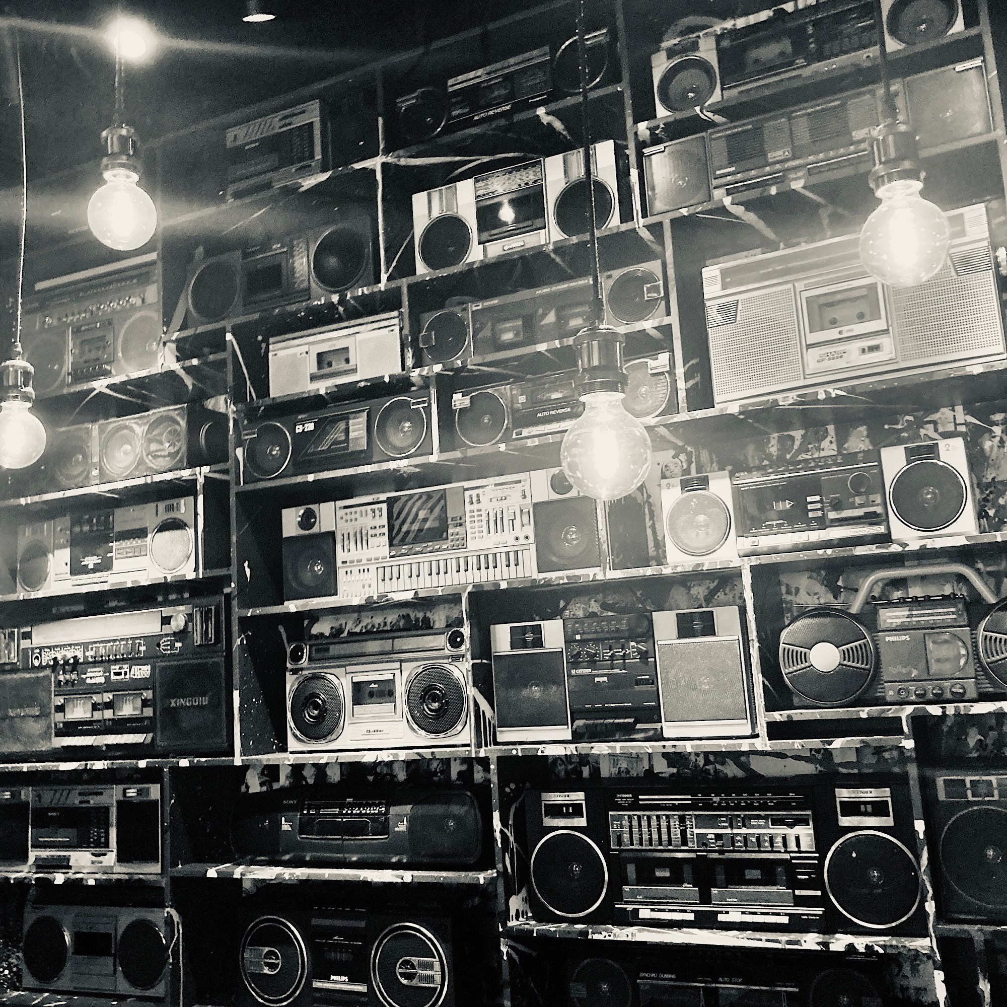 Rows of vintage cassette players and radios cover a wall between hanging bare bulbs.