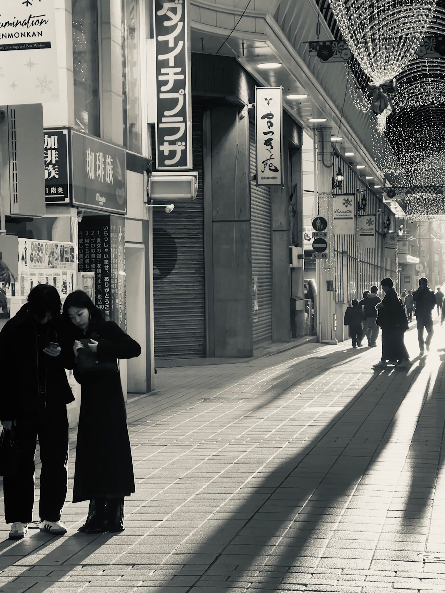 Pedestrians cross a covered shopping arcade under Japanese signs and long shadows.