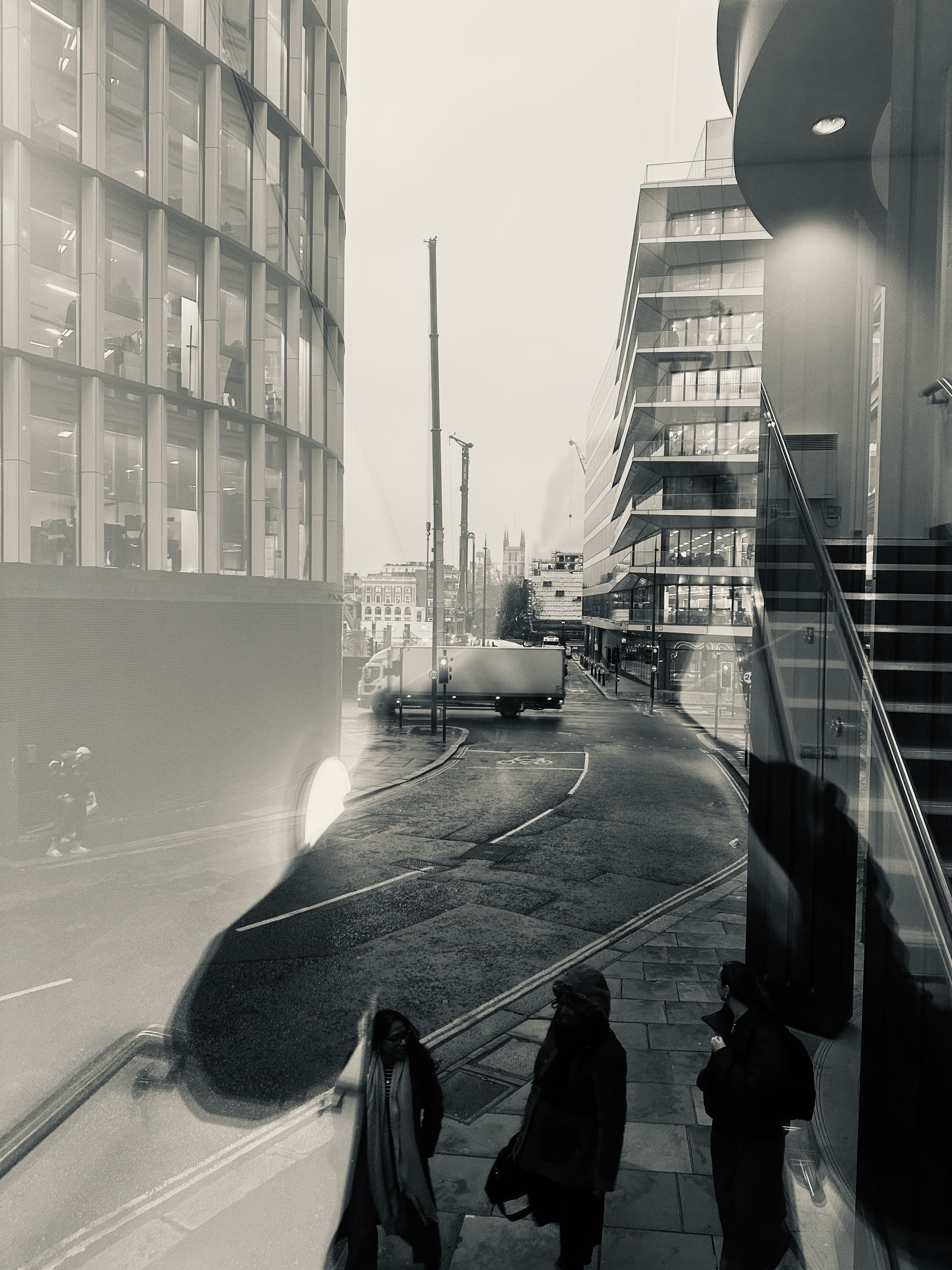 Pedestrians and a lorry layered in glass reflections between city buildings and cranes.
