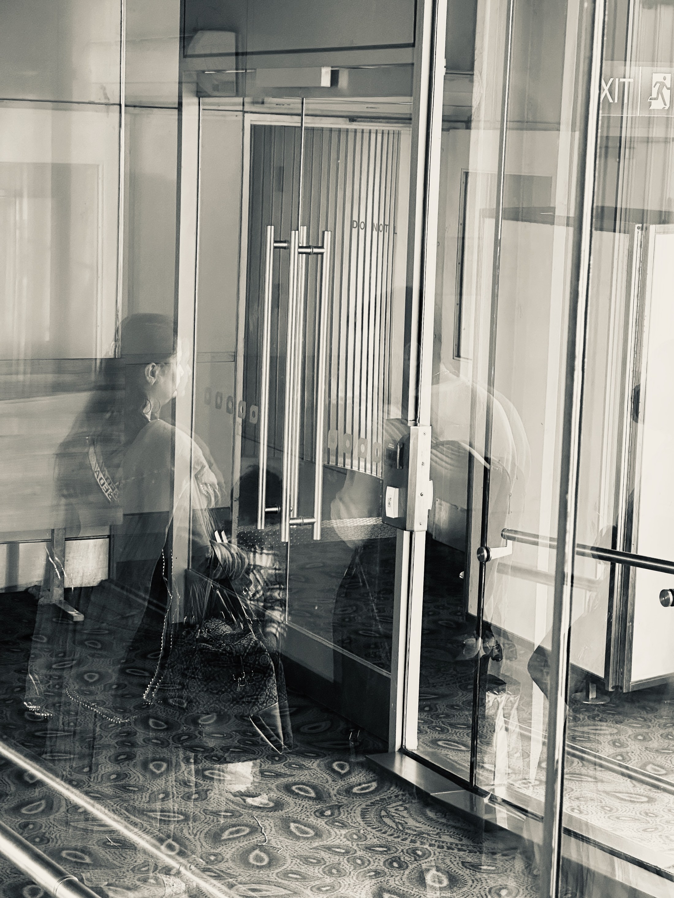 A blurred figure is split by reflections and the vertical frames of glass entrance doors.