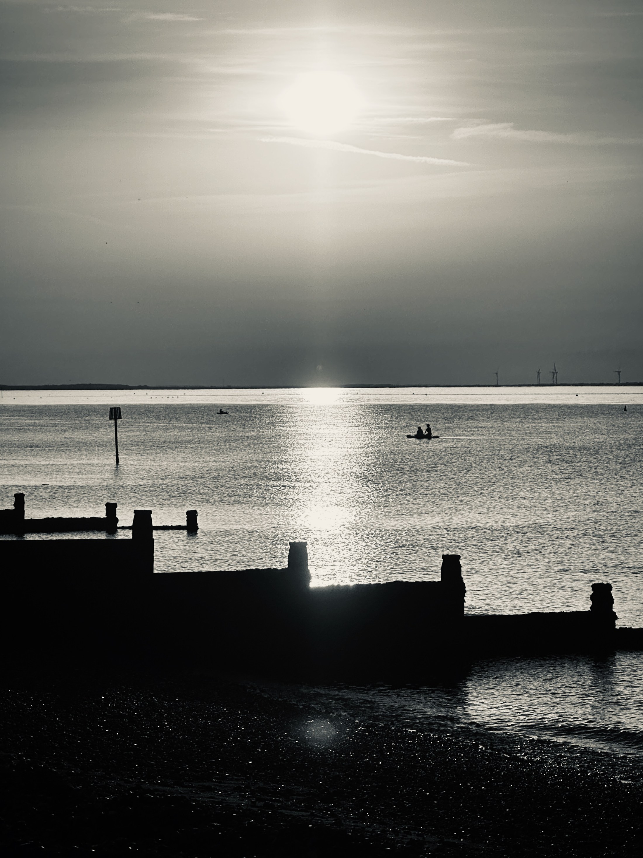 A vertical path of sunlight across the sea beyond a dark promenade.