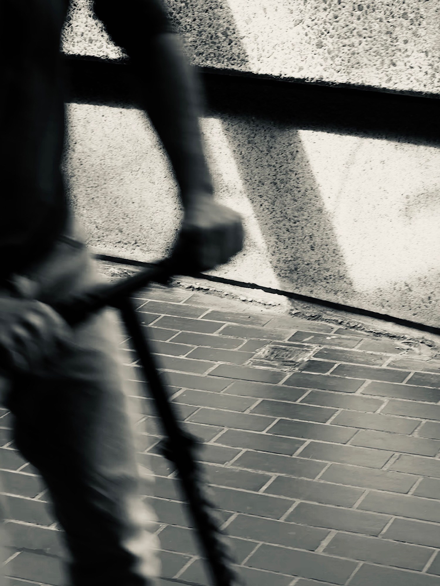 A cyclist blurred in motion crosses hard diagonal shadows on a pale wall.