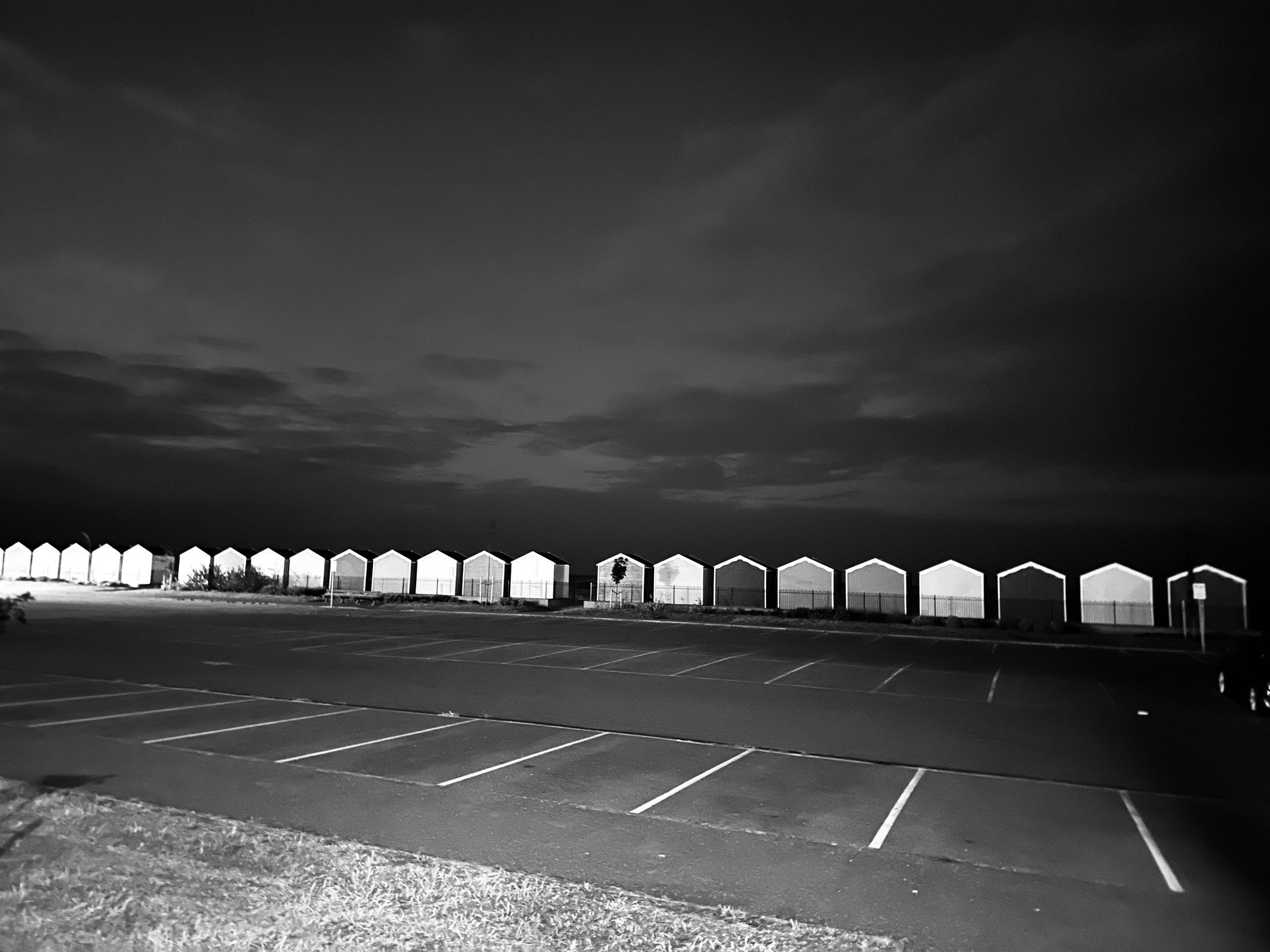 A row of bright huts beneath a dark sky and an empty car park.