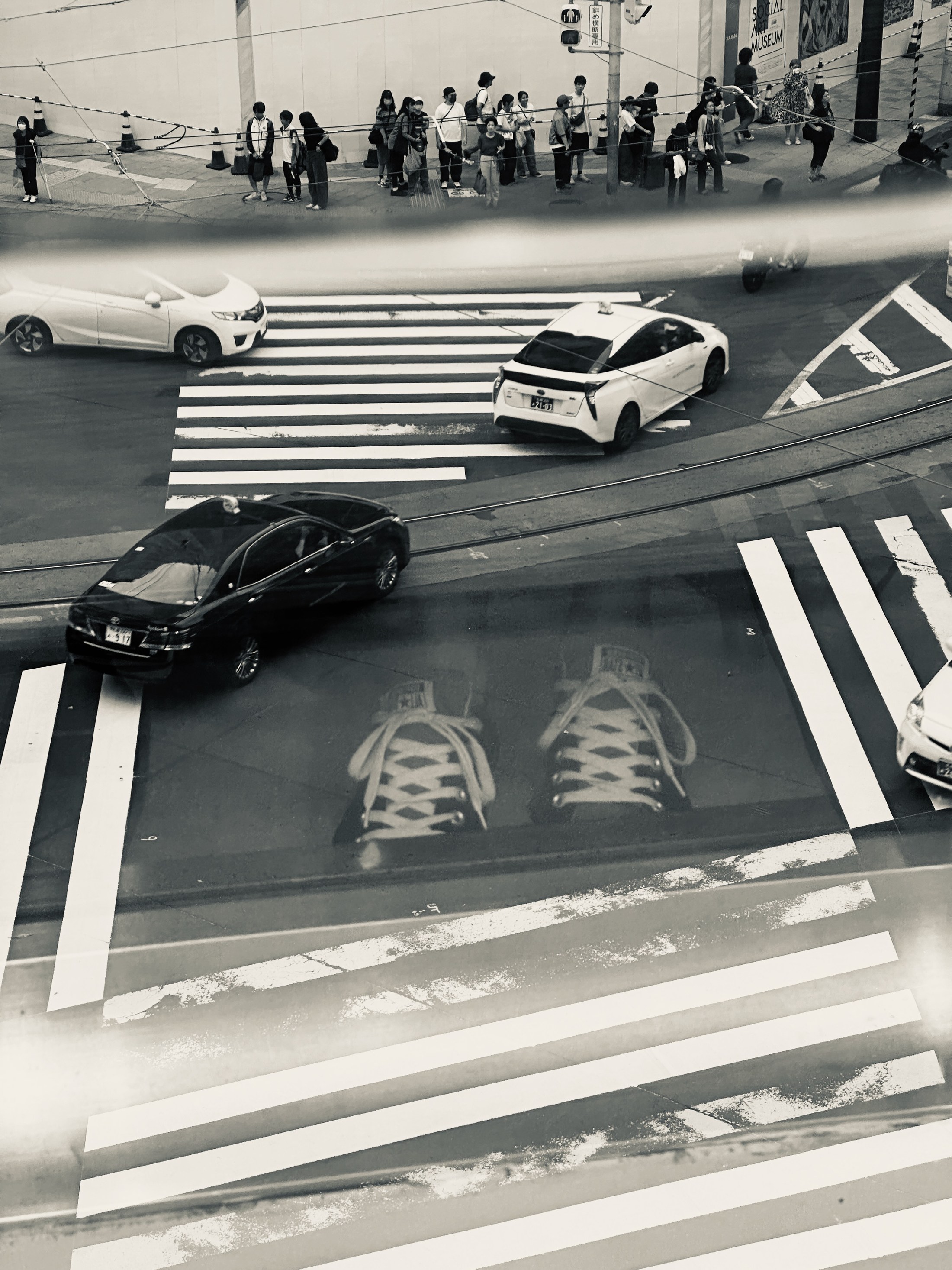 An elevated view of a zebra crossing, traffic and a waiting crowd, overlaid by reflected shoes.