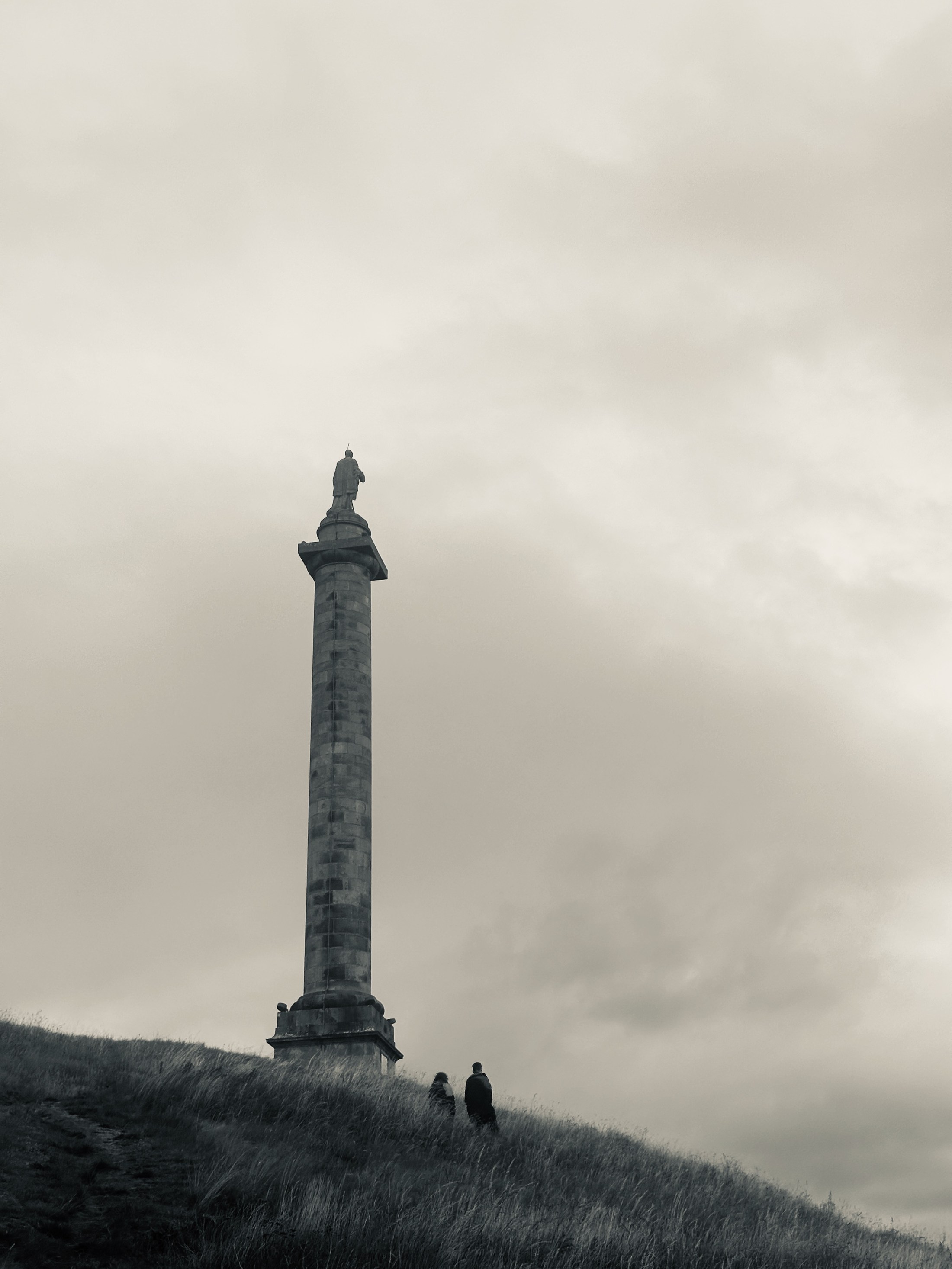 A tall stone column rises into a broad clouded sky above two small figures walking on a sloping grassy hill.