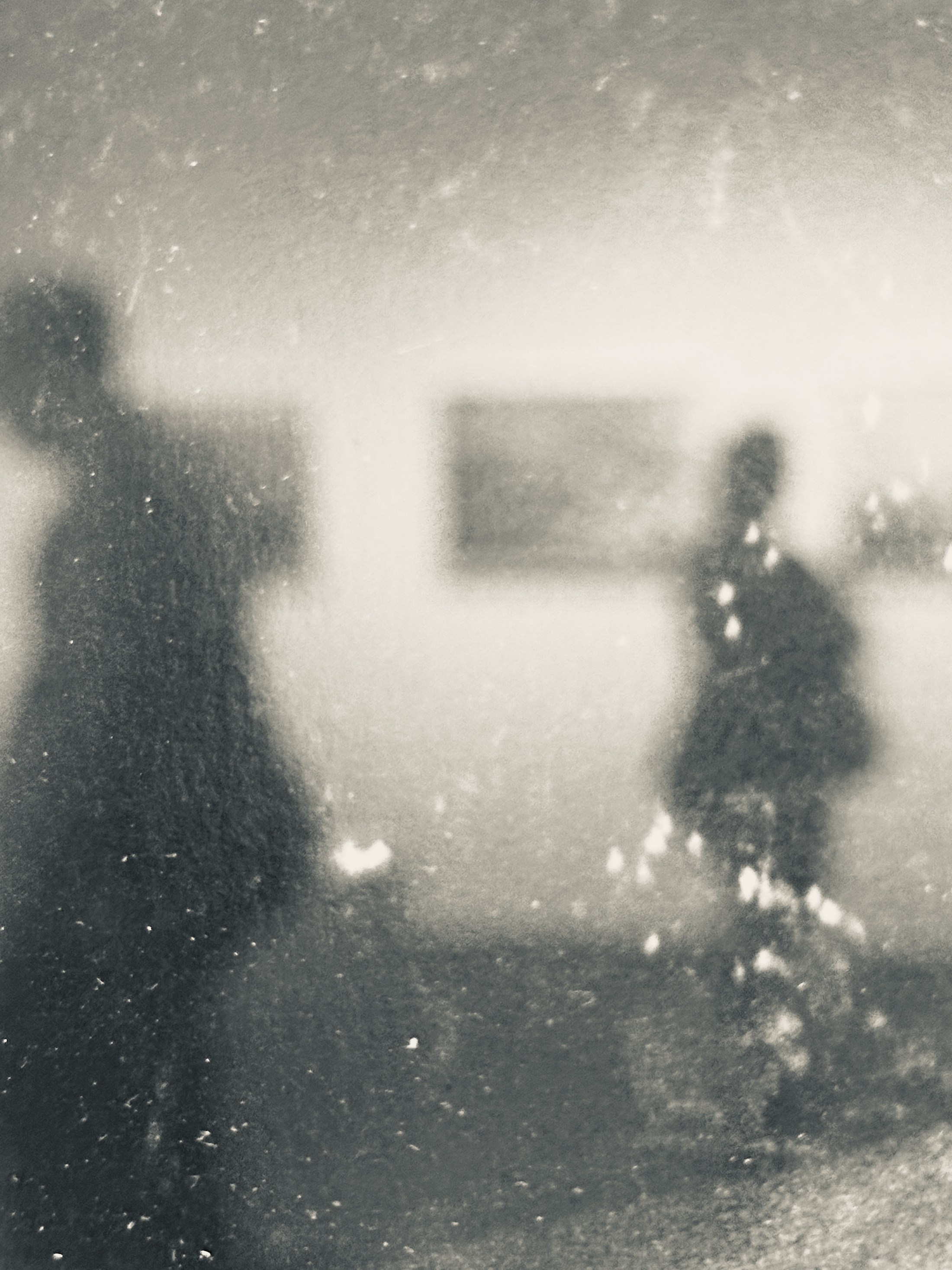 Two blurred silhouettes at different scales seen through dirty glass, with light and surface marks dissolving their outlines.